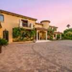 A stucco home with a tile roof, arched entry and a wide paver driveway at dusk.