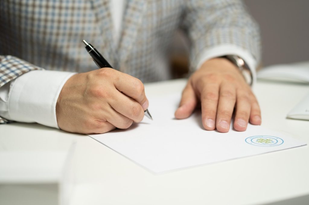 A person signing a document with a pen, with papers and a contract visible on the surface.