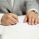 A person signing a document with a pen, with papers and a contract visible on the surface.